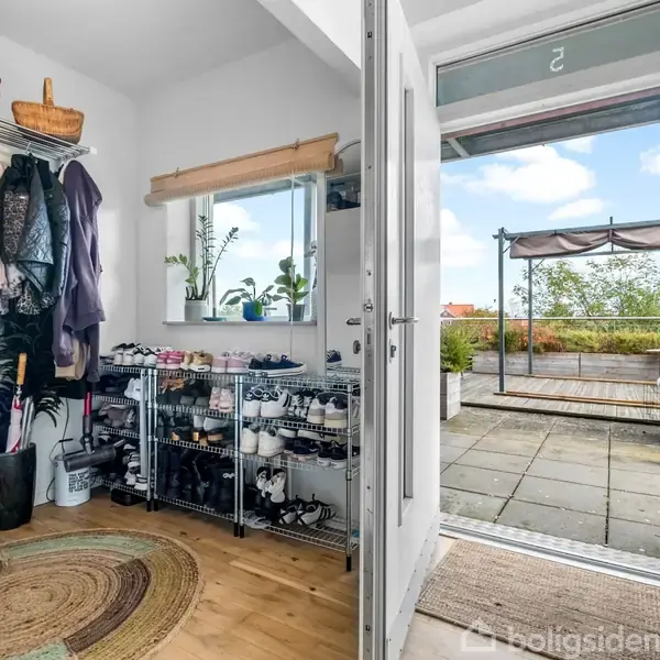 Entré med jakker hængende på knager, sko stablet i reoler og en rund tæppe på trægulvet. Døren står åben mod en terrasse med udsigt til himmel og planter.