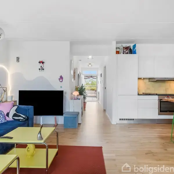 Sofa med farverige puder står til venstre, foran et tv. Køkken med hvidt inventar og barstole til højre. Lys træparket dækker gulvet. I baggrunden ses en gang.