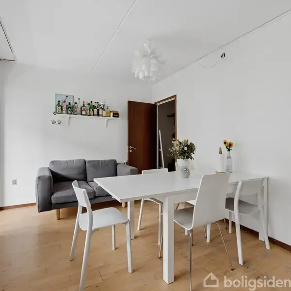 Hvidt spisebord med fire stole står i en lys stue. En grå sofa og en hylde med flasker er i baggrunden. Plantepotter pynt vindueskarmen.