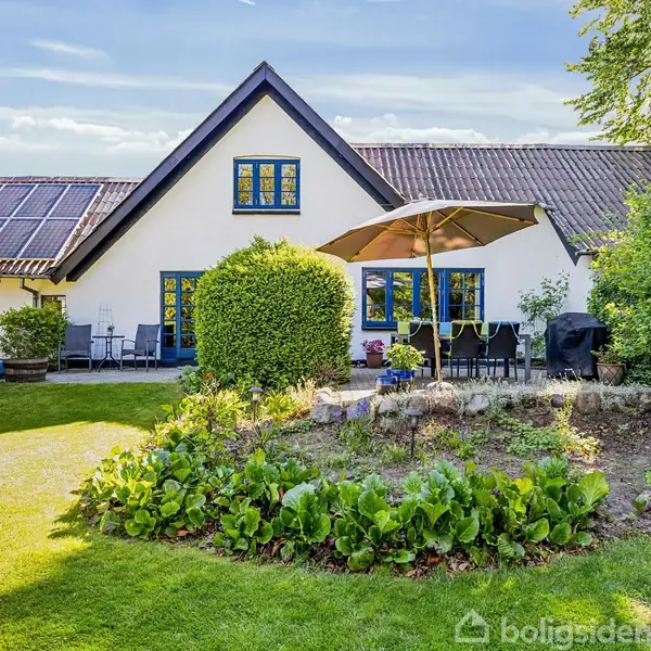 Hvid hus med blå vinduer, havebord med parasol i forgrunden, omgivet af grøn have og buske. Terrasse til venstre, blå himmel og sollys rammer scenen.
