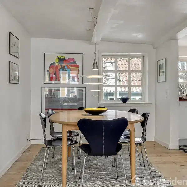 Rundt spisebord med sorte stole står i et lyst rum med hvidmalede vægge. Bordet er dækket med en skål, omgivet af kunstværker på væggene og vinduer med lysindfald.