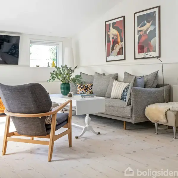 En stue med en grå sofa fyldt med puder, ved siden af en gulvlampe og skindbetrukket skammel. Et træmøbel står under et vindue, mens malerier hænger på væggen.