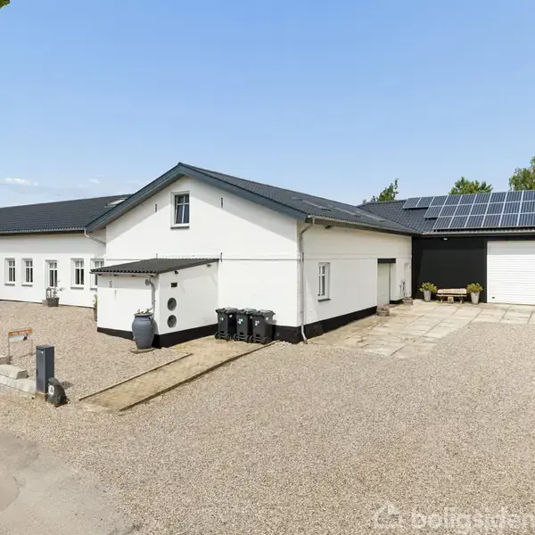 En hvid bygning med sort tag udstyret med solpaneler, placeret i en grusbelagt gårdsplads; omkranset af grønne træer under en blå himmel.