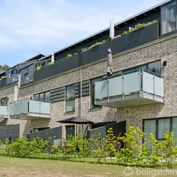 Rækkehuse med glasbalkoner strækker sig langs en grøn have med små buske. Foran bygningerne står parasoller mellem terrasser med glasdøre. Himlen er klar og blå.
