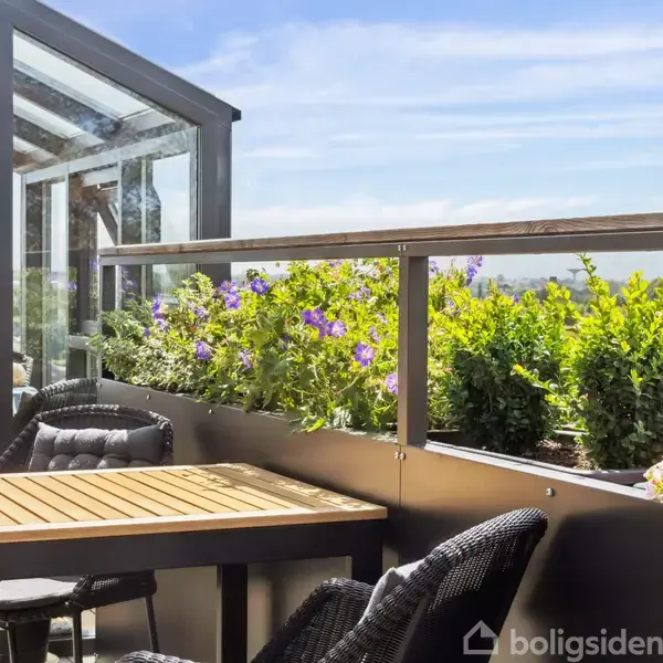 Terrassemøbler står på en solbeskinnet altan med glasværn. Blomster og grønne planter pryder rækværket. I baggrunden ses en landskabsudsigt med træer og blå himmel.