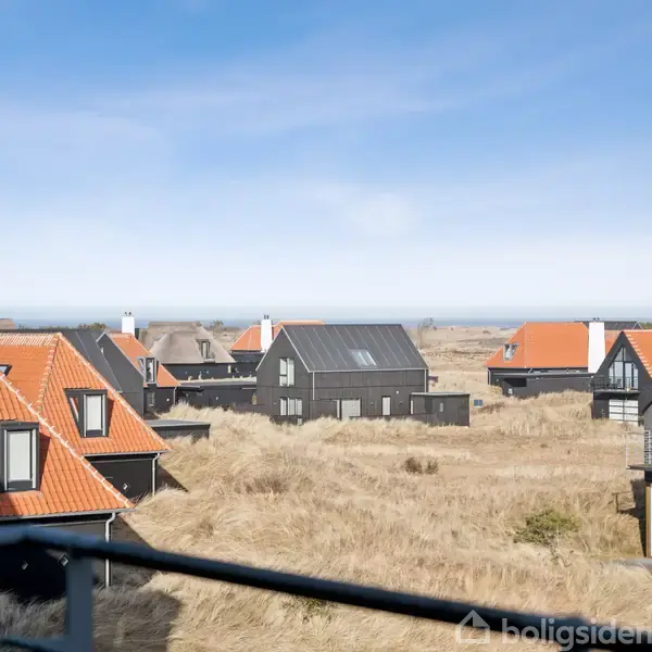 Huse med sorte facader og orange tage står spredt i et kystnært landskab med græsbevoksede klitter, under en blå himmel.