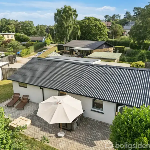 En hvid, en-etagers hus med sort tag, omgivet af grønne træer. Der er en terrasse med liggestole og en stor, hvid parasol, der skaber en afslappet atmosfære.