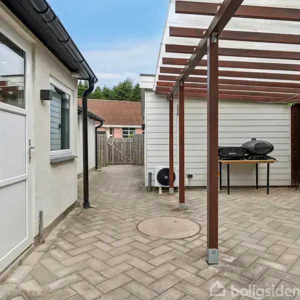 Terrasse med belægningssten og halvtag. Til højre står en grill på et bord tæt op ad en hvid bygning med træbeklædning. Til venstre ses en hvid dør og vindue.