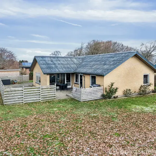 Et gult murstenshus med sort tag står stille omkranset af en græsplæne dækket af efterårsblade. Foran huset er en terrasse indhegnet af et træhegn.