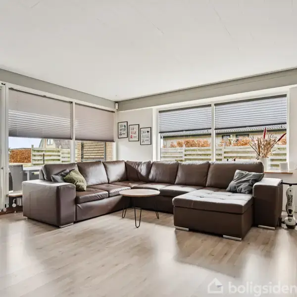 Stor brun hjørnesofa står i en lys stue med trægulve, vendt mod vinduer med udsigt til haven. En reol med bøger og pynt står i hjørnet.