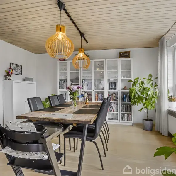Spisebord med sorte stole står i spisestuen, oplyst af hængelamper. Skab med glaslåger og planter i potter omkranser rummet. Rummet er lyst med stort vindue til højre.