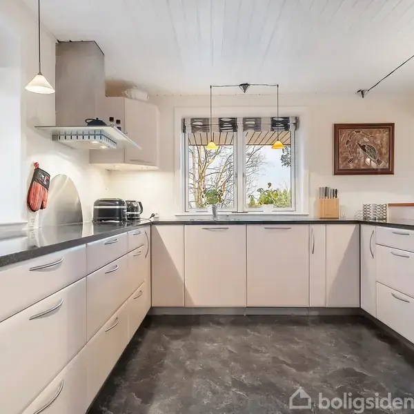 Moderne køkken med hvide skabe og skuffe. Køkkenet har sorte bordplader, indbygget ovn og emhætte. To vinduer bringer naturligt lys ind. Gulvet er mørkt, og der hænger kunst på væggen.