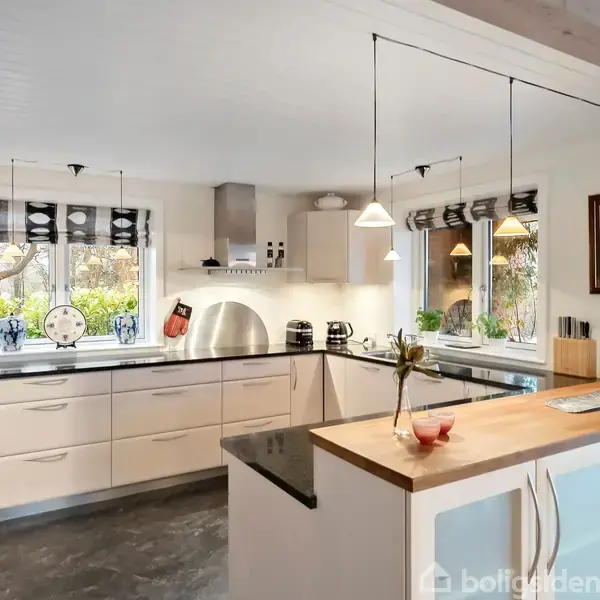 Et moderne køkken med hvide skabe og en sort bordplade. Et træskåret køkkenbord står centralt. Store vinduer giver udsyn til naturen. Hængelamper tilfører lys.