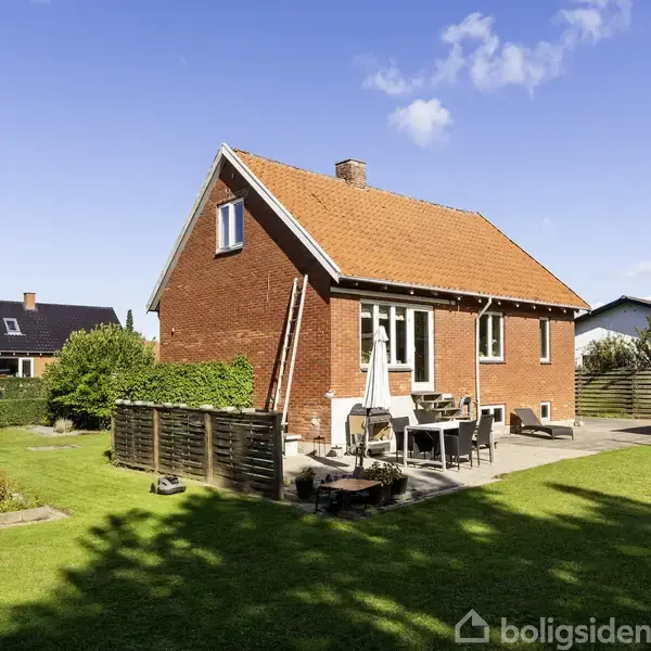 Rødt murstenshus med tegltag står i en have. Solsenge og havestole placeret på terrasse. Grønt græs omgiver huset, mens andre huse ses i baggrunden.