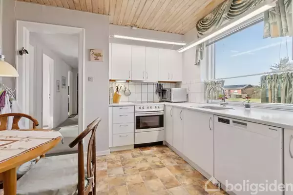 Et køkken med hvide skabe har en komfur, en mikrobølgeovn og en opvaskemaskine. Et rundt spisebord med stole står til venstre. Vindue viser udenfor landskab. Skilt: "Sicilians Stue."