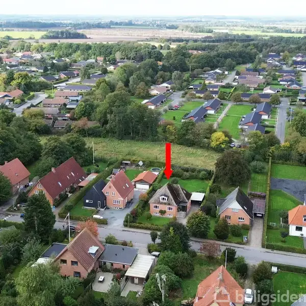 Hus under en rød pil, omgivet af andre huse, træer og haver i en lille by. Landskabet er grønt og frodigt med marker i baggrunden.