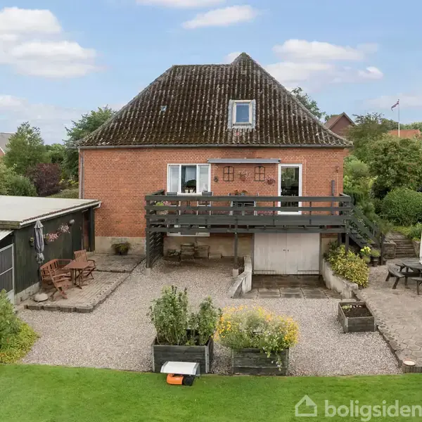 Et rødt murstenshus med en stor terrasse og udendørs møbler i en have omkranset af buske. Græsplæne og terrasseområder skaber en hyggelig udendørs atmosfære.