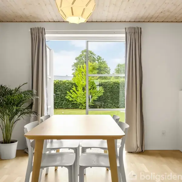 Et spisebord med fire stole står i et lyst rum med en stor glasdør. Udenfor er en grøn have med buske og træer. Indendørs er der planter og lyse gardiner.