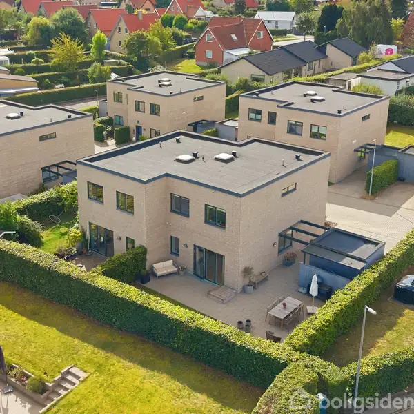 Flere moderne, kubiske huse med fladt tag er placeret i en forstad. Et centralt hus har en velplejet have og terrasse med møbler. Omkringliggende huse og grønne områder ses i baggrunden.