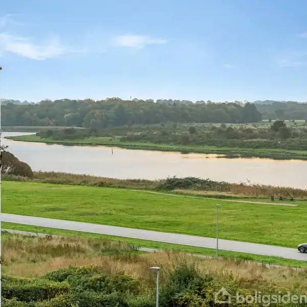 En flod løber stille gennem et grønt, frodigt landskab. Træer omkranser området, mens en asfalteret vej snor sig langs flodens bredder under en klar, blå himmel.