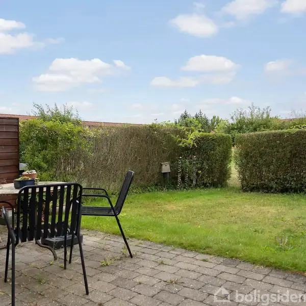 Et terrasseområde med to stole og et bord, omgivet af en plæne og hæk, under en blå himmel med få skyer.