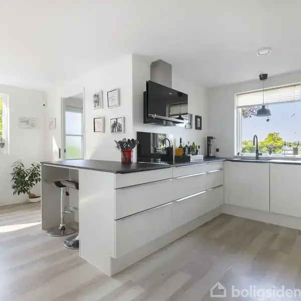 Et moderne køkken med hvide skabe og en sort bordplade. Barstol står ved køkkenøen. Store vinduer giver lys og udsigt til have. Plante og indrammede billeder ses.