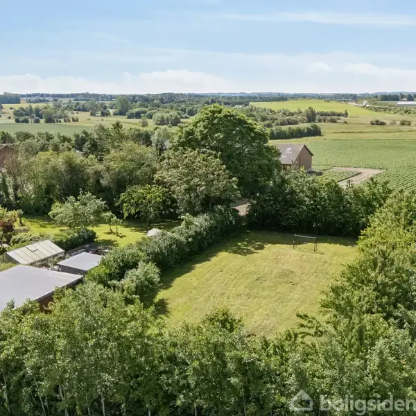 Grønt jordareal er omkranset af træer og buske. Det ligger i et landligt område med marker og skove, under en klar blå himmel.