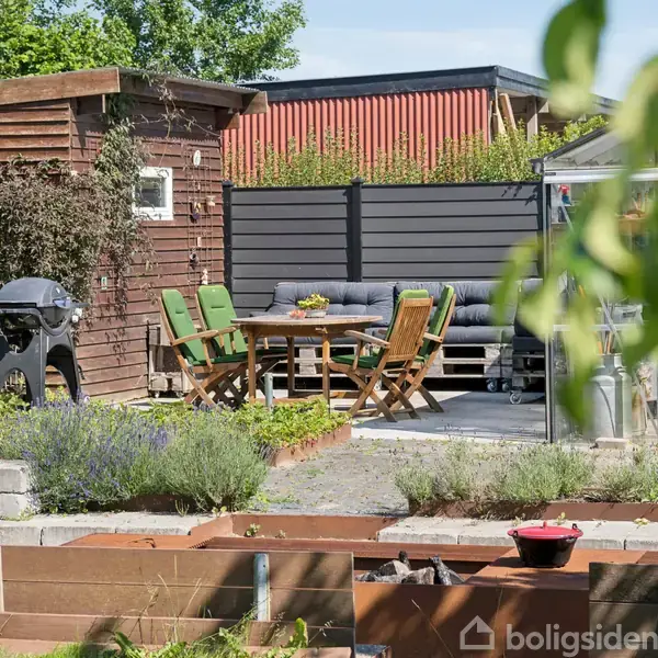 Haveområde med træmøbler og grill i midten, omgivet af et skur og planter. I baggrunden ses hegn og en bygning. Grønt løv er delvist i forgrunden.