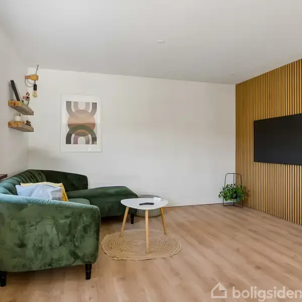 Grøn sofa står i stuen, accenteret af et rundt bord og vægkunst. Rummet har en trælistevæg med en fladskærm og er lyst med et stort vindue til venstre.