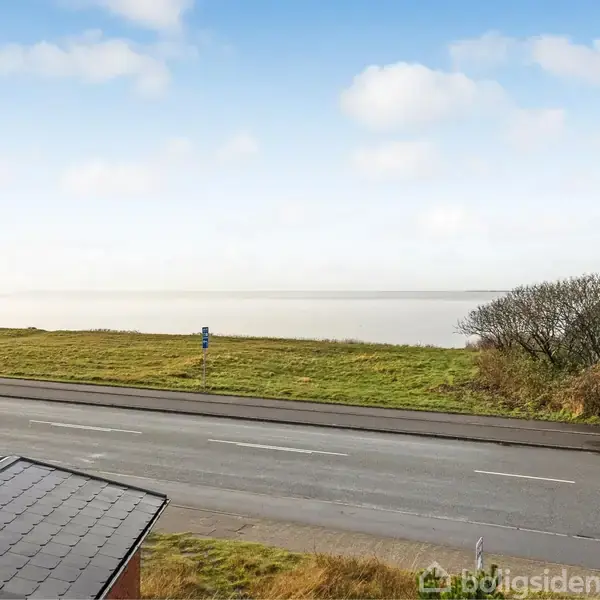 Vejen strækker sig forbi et hustag i forgrunden mod en græsbeklædt kystlinje; himlen er let overskyet med spredte skyer.
