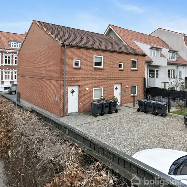 Rød murstensbygning står stille på en grusbelagt parkeringsplads, omgivet af hække og naboejendomme. Flere skraldespande er opstillet ved siden af indgangsdørene, og biler parkerer tæt ved.