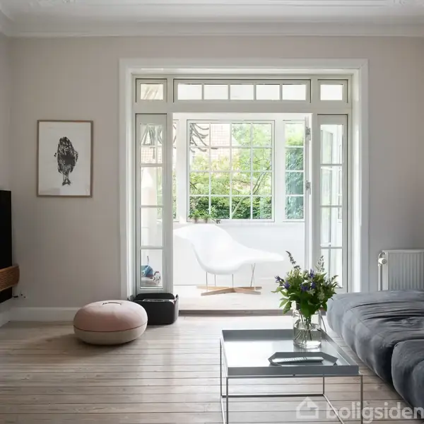Fladskærms-tv hænger på væggen, mens en stor grå sofa står i opholdsstue med trægulv; en glasdør åbner mod en lys veranda med grøn udsigt.