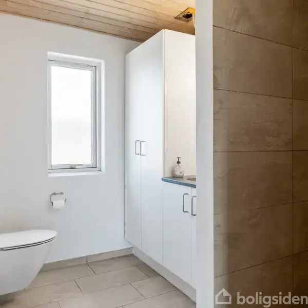 Toilet placeret ved siden af et smalt vindue, med skab og vask til højre. Flisebelagt bruseniche ses i højre del af billedet. Lys, moderne badeværelse med neutralt farvevalg.