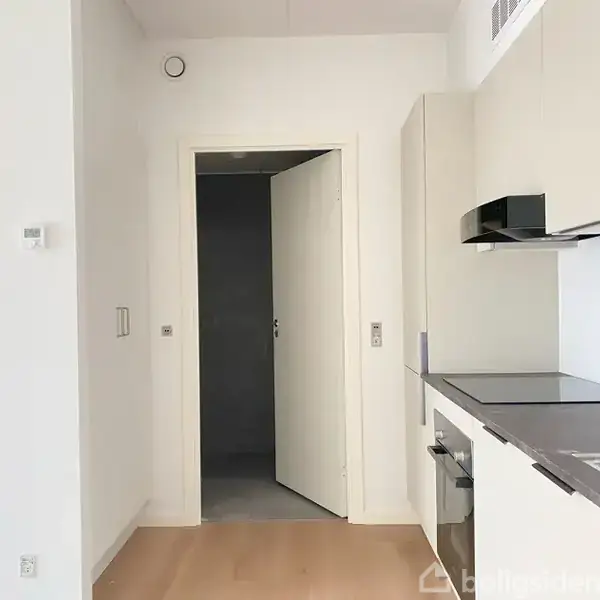 Et køkken med hvide skabe og indbygget ovn. Der er en åben dør til en mørkere gang. Gulvet er lyst træ, og der er minimal dekoration.