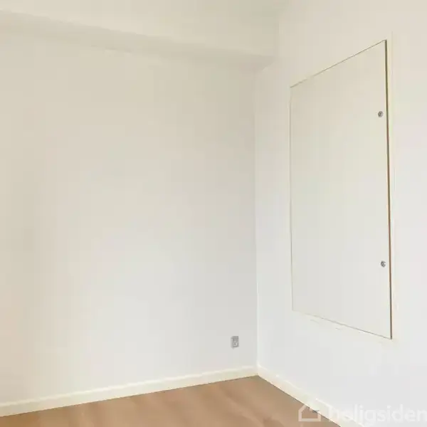Et hvidt klædeskab med to døre står i et lyst rum. Skabet er placeret ved en hvid væg med en dør og en åbning på højre side.