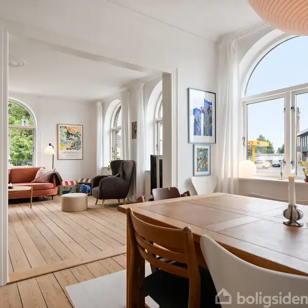 Et spisestuebord med stole står i forgrunden, mens en stue med sofa og lænestol er synlig i baggrunden, omgivet af store vinduer og lyse vægge.