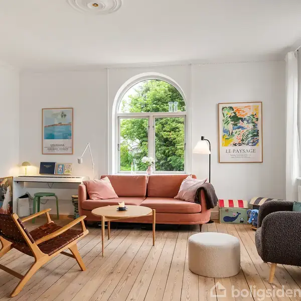 Stue med sofa og lænestole, placeret omkring et rundt sofabord. Der er plakater på væggene og store vinduer, der giver udsigt til grønne omgivelser.