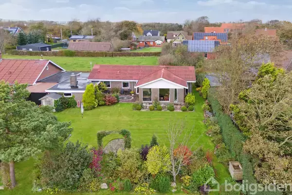 Huset med rødt tag står på en velplejet græsplæne omgivet af farverige buske og træer i en landlig boligkvarter. I baggrunden ses flere huse og grønne marker.