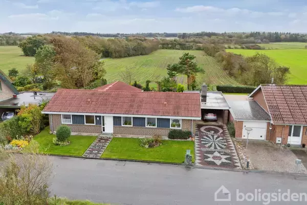 Hus med rødt tegltag og brun murstensfacade, omgivet af velholdt have og indkørsel med dekorativt flisemønster. Beliggende i landligt område med grønne marker og træer i baggrunden.