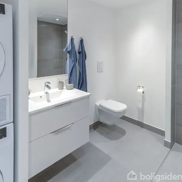 En moderne badeværelse med en vaskemaskine og tørretumbler stablet ved siden af en håndvask. Et væghængt toilet og bruseniche fuldfører det lyse rum.