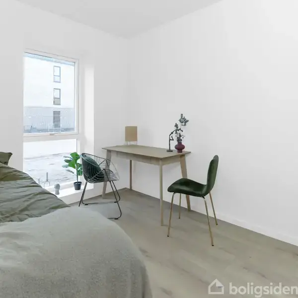 Et enkelt indrettet værelse med en seng i venstre side. Et skrivebord med en lampe og plante står op ad vinduet i midten. Rummet er lyst og minimalistisk.