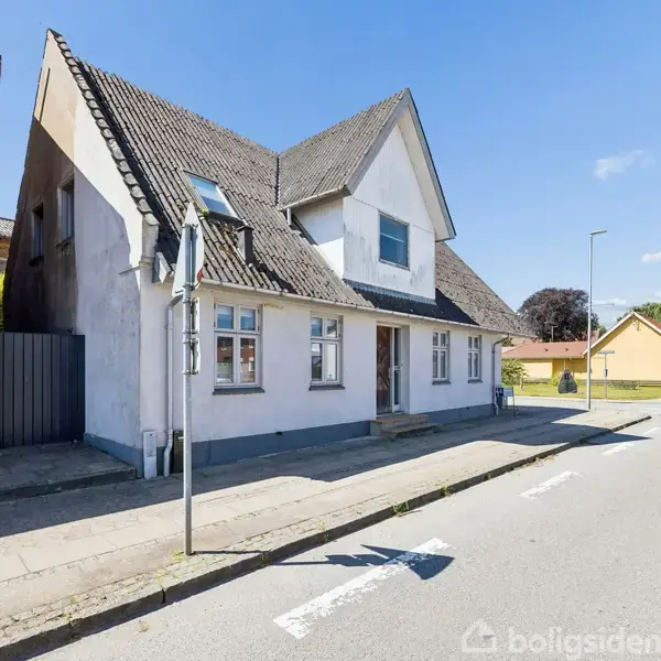 Hvidmalet to-etagers hus med spidst tag står ved siden af en rød murstensbygning. Solrig gade med fortov og få skyer på den blå himmel.