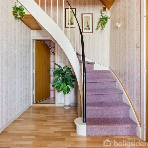 Trappe med lyserød tæppebelægning, der fører opad, omgivet af lyse vægge og træpaneler. Grønne planter og indrammede billeder hænger på væggene i en entrélignende passage.