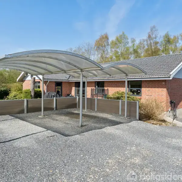 Carport står tom på grusvej foran rødt murstenshus, omgivet af træer og buske under blå himmel.