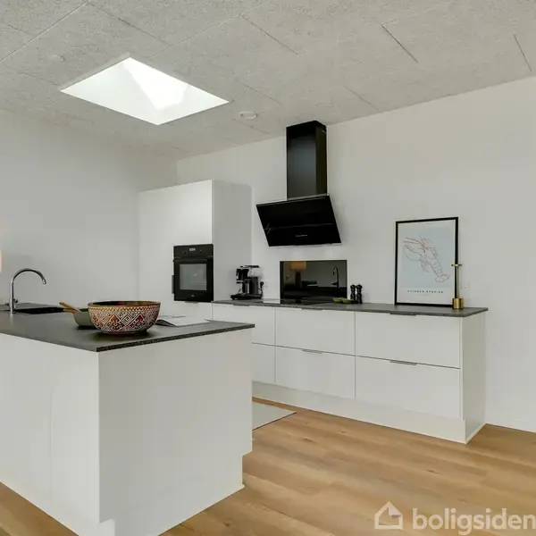 Køkken-alrum med hvide skabe og sort bordplade; spisebord med stole i hjørne; lys fra stort vindue; træplankegulv og loft med spotlys.