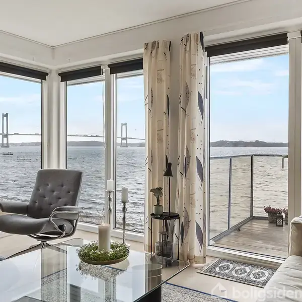 Stue med moderne møbler; en lænestol og glasbord står ved store vinduer, der åbner op til en balkon med udsigt over vandet og en bro i horisonten.