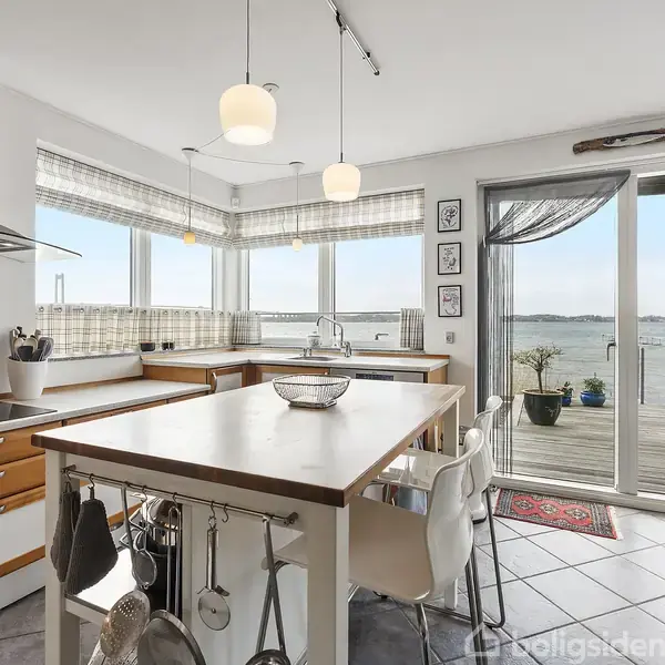 Et lyst køkken med træbord og barstole, omgivet af lyse skabslåger. Store vinduer og en skydedør åbner mod en terrasse med udsigt over havet i baggrunden.