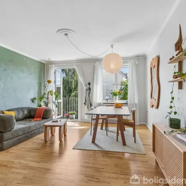 Stuen har en grå sofa med puder, et sofabord og spisebord. Grønne planter står ved vinduerne, som lader sollys komme ind. Trægulv og hvide vægge med grøn accent.