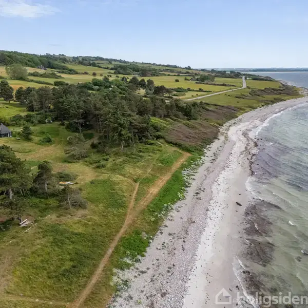 Kyststrækning langs sandstrand, hvor bølgerne slår mod bredden. Skovklædt område og marker strækker sig langs kysten. En vej følger landskabets konturer i baggrunden.