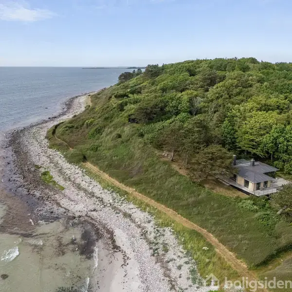 Hus ligger fredfyldt ved en træfyldt skrænt ud til en stenstrand. Skovklædt bakke omkranser huset, og havet strækker sig i horisonten, hvilket skaber en naturskøn kystudsigt.